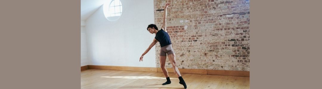 a male ballet dancer posing against a sunlight brick wall wwearing imperfect pointes brown dance shorts and black top