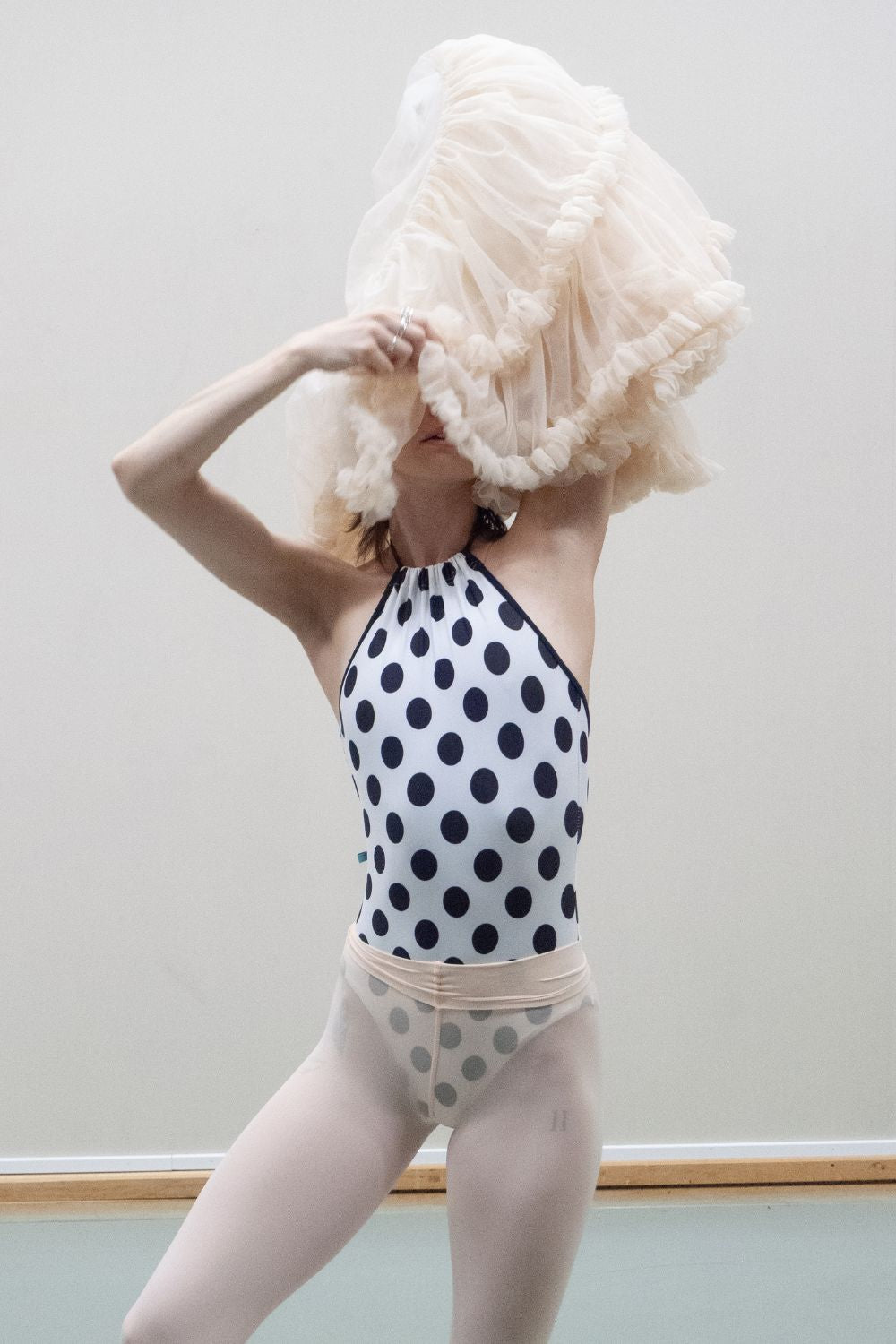 A professional ballet dancer wearing the Imperfect Pointes Eastbourne tie-neck leotard in white with black polka dots, styled with a tutu and tights over leotard in a ballet studio