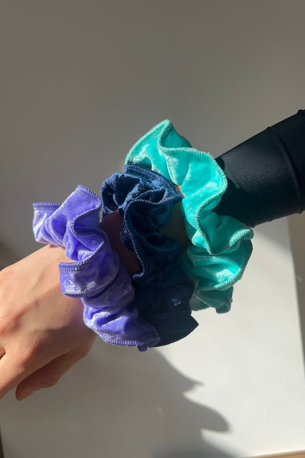 person wearing 3 crushed velvet scrunchies on wrist, one light purple, one navy blue and one mint green. 