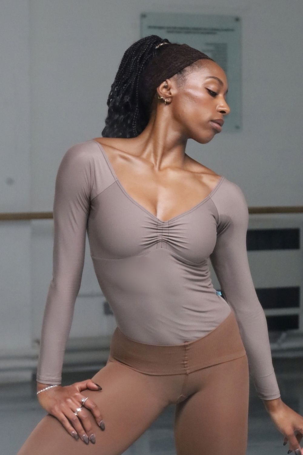 A black ballerina posing in a dance studio wearing a taupe long-sleeve ruched leotard and matching skin tone tights from imperfect pointes