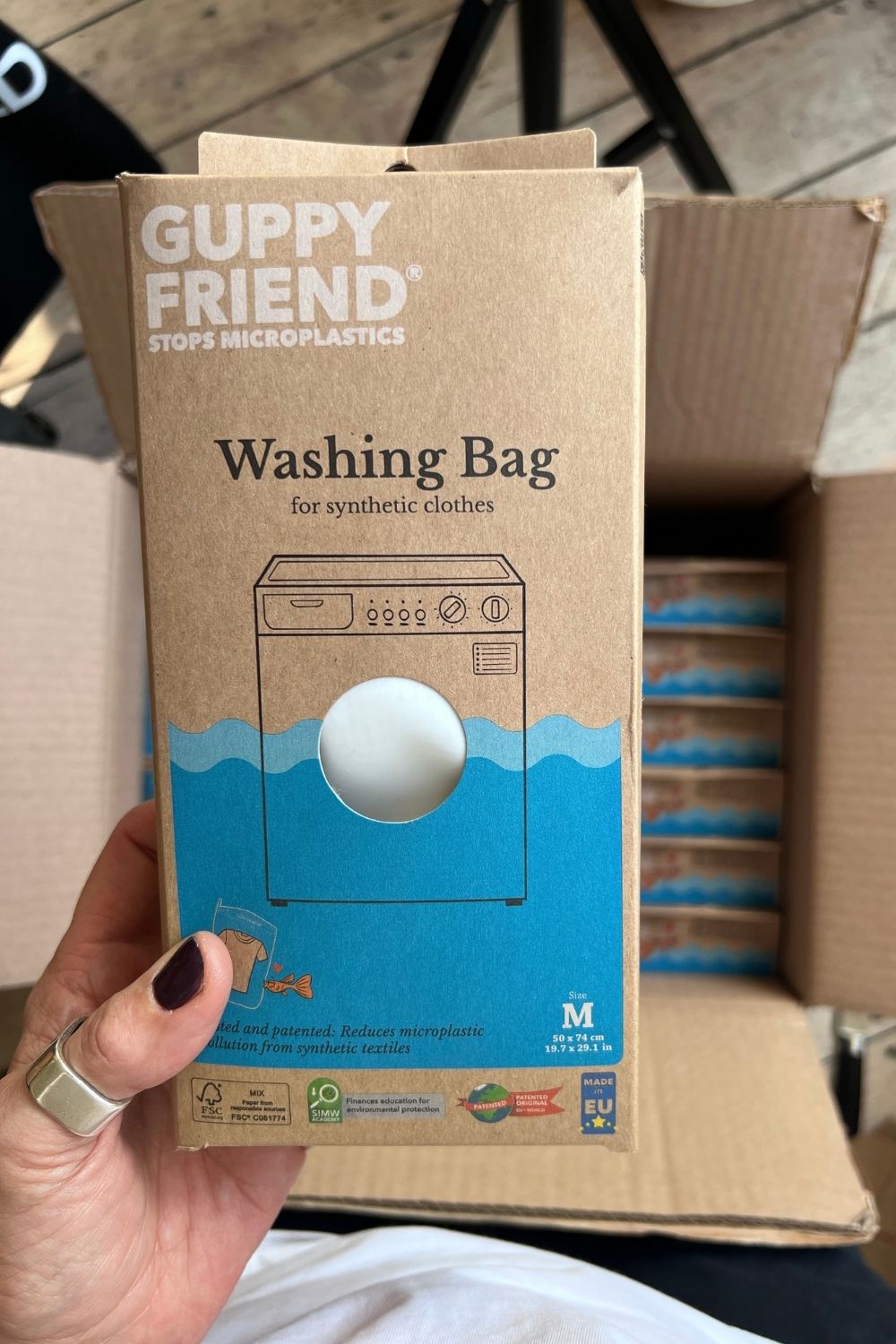 Hand holding a GUPPYFRIEND washing bag box in front of an open carton of packaged bags