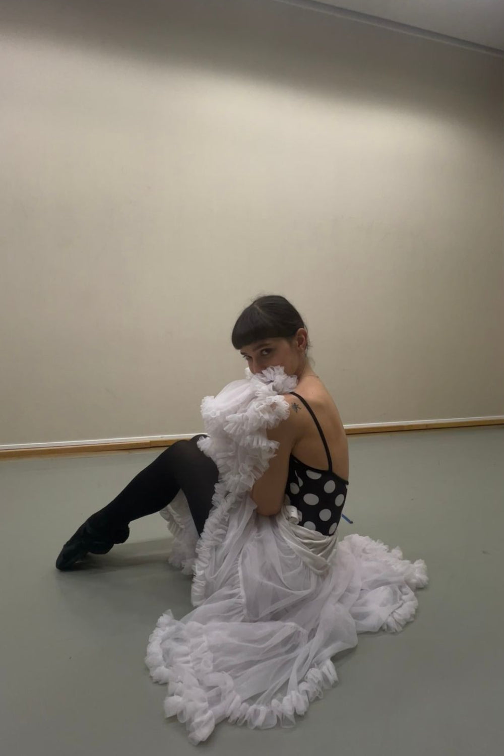 A professional ballerina wearing the Imperfect Pointes Cali Camisole polka dot leotard – seated in a dance studio with a white feather skirt