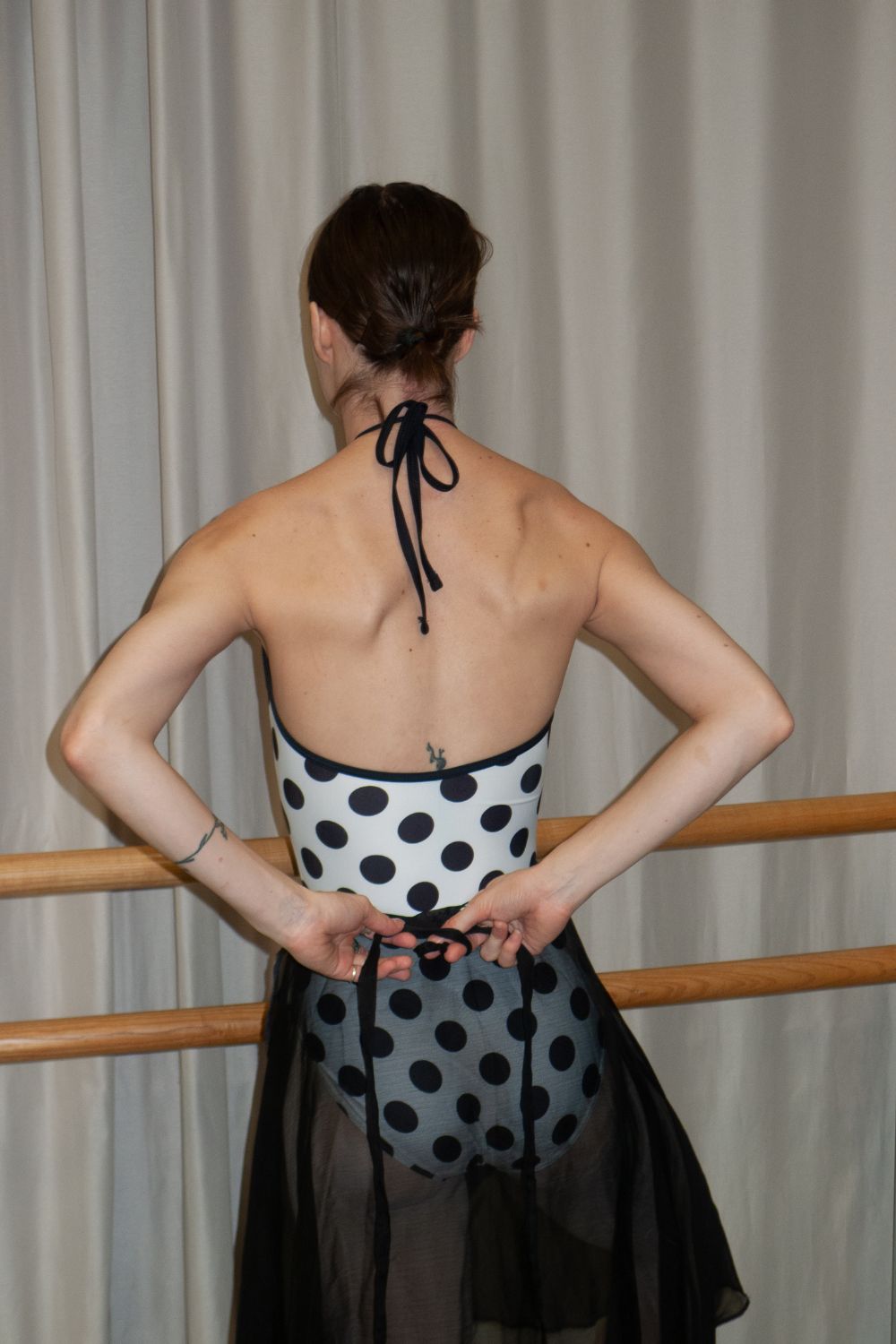 A professional ballet dancer wearing the Imperfect Pointes Eastbourne tie-neck ballet leotard – back view showing the open back and tie-neck detail with a black mesh skirt against the ballet studio wall.