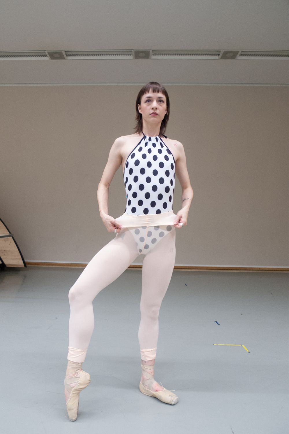 A professional ballet dancer wearing the Imperfect Pointes Eastbourne halter neck leotard – full length shot showing the white with black polka dot print, turning down the waistband of their tights