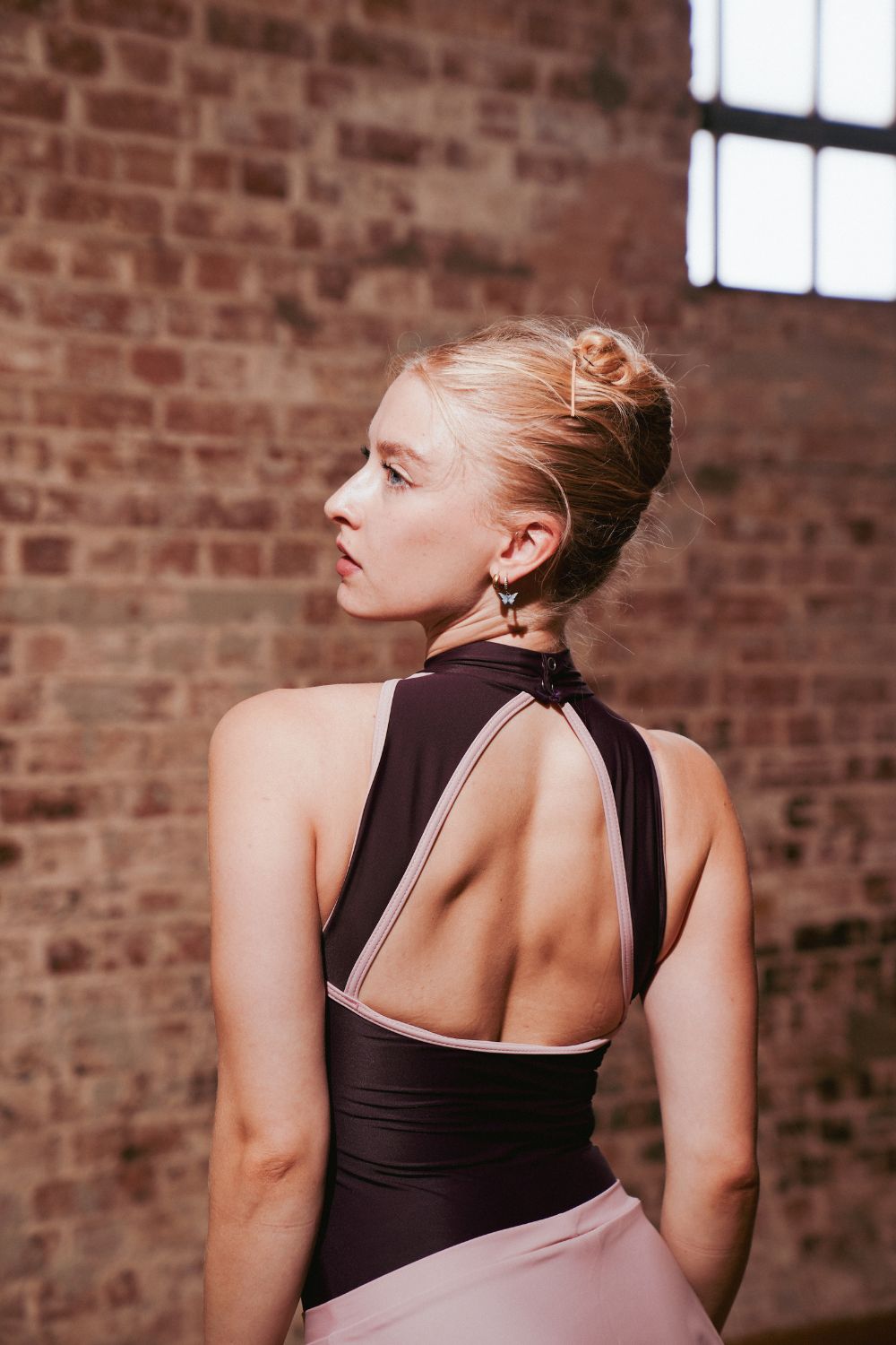 ballet dancer with blonde hair in a fench twist wearing a newly launched Imperfect Pointes halterneck leotard leotard in cherry lacquer . viewed from the back against a brick wall 